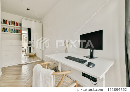 A sleek and modern home office featuring a minimalist desk, computer, and stylish bookshelf in a bright, airy room with warm wooden flooring. 123894862