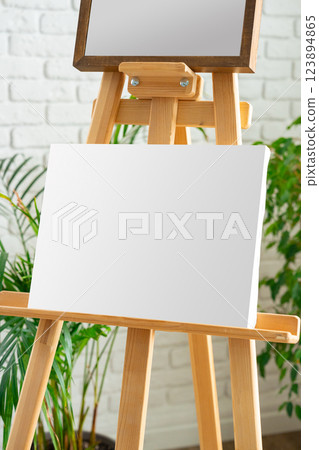 Blank canvas on wooden easel beside green plants in a bright indoor space perfect for artistic inspiration and creativity Blank canvas on wooden easel beside green plants in a bright indoor space perfect for artistic inspiration and creativity 123894865