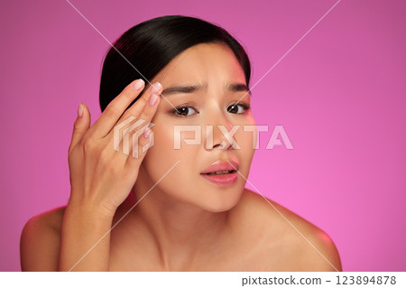 Young woman applying skincare product on her face in front of a vibrant pink background 123894878