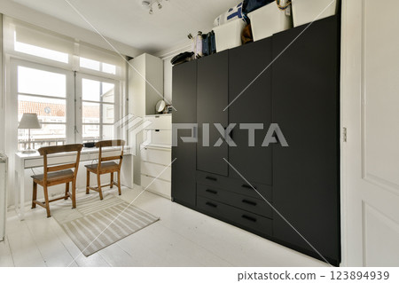 A sleek, modern bedroom featuring a minimalist design, characterized by a dark wardrobe, wooden chairs, and abundant natural light from large windows. A sleek, modern bedroom featuring a minimalist design, characterized by a dark wardrobe, wooden chairs, and abundant natural light from large windows. 123894939