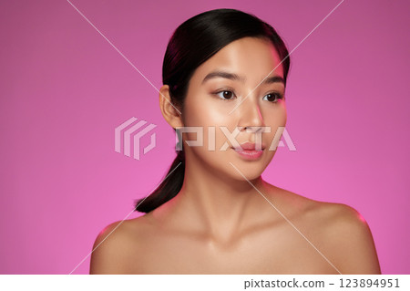 Woman with smooth skin posing against a vibrant pink background in a studio setting, showcasing makeup artistry and beauty 123894951
