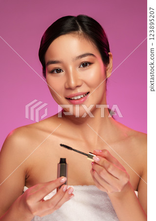 Woman applying mascara while smiling in a beauty setting with soft pink background Woman applying mascara while smiling in a beauty setting with soft pink background 123895207