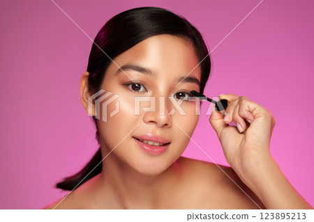 Young woman applying mascara against a vibrant pink background in a beauty setting 123895213