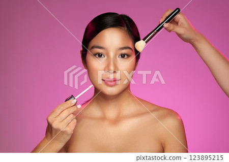 Makeup artist applies lip gloss and blush to model with pink backdrop during studio photoshoot 123895215