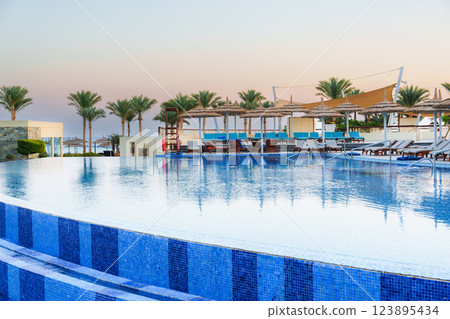 Luxury resort pool area at sunset with palm trees and lounge chairs in a tranquil setting by the sea 123895434