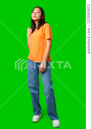 Young woman poses confidently in an orange shirt and jeans against a bright green background highlighting her stylish presence 123895623