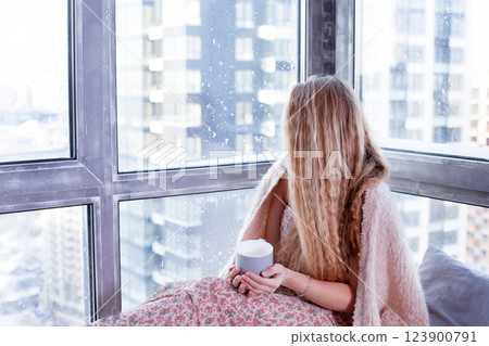 Rest at home by the window. A woman sits and drinks coffee. Long-haired beautiful blonde girl. Comfort in winter. Cozy morning mood Rest at home by the window. A woman sits and drinks coffee. Long-haired beautiful blonde girl. Comfort in winter. Cozy morning mood 123900791