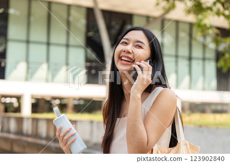 Sustainable lifestyle. Young woman laughing on the phone while showcasing her reusable cup. 123902840