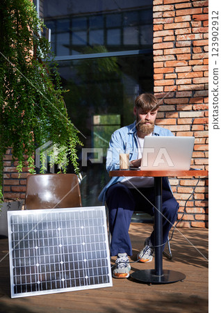 Man works on laptop at outdoor round table, with glass of iced coffee. Solar panel nearby, emphasizing sustainable, modern eco-friendly workspace that combines technology and sustainability. 123902912