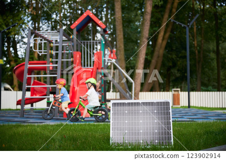 Solar panel for charging in foreground on grassy area of playground. Two children wearing helmets ride balance bikes near vibrant red play structures, surrounded by tall trees and fenced boundary. Solar panel for charging in foreground on grassy area of playground. Two children wearing helmets ride balance bikes near vibrant red play structures, surrounded by tall trees and fenced boundary. 123902914