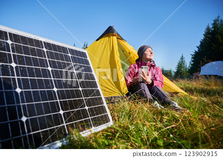 Happy woman sits in front of yellow tent, charging her phone by using solar panel, highlighting use of renewable energy and eco-friendly camping. On background lush green trees and clear blue sky. 123902915
