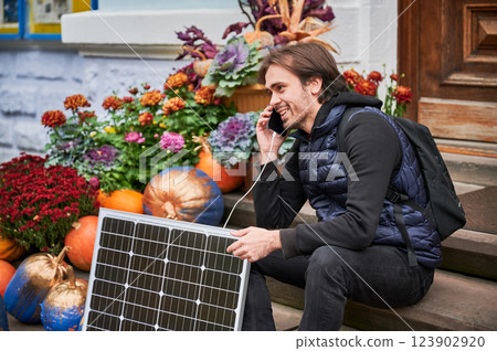 Handsome man talks on smartphone connected to photovoltaic solar panel. Integration of sustainable renewable energy into everyday life, demonstrating practical use of solar power for charging devices. Handsome man talks on smartphone connected to photovoltaic solar panel. Integration of sustainable renewable energy into everyday life, demonstrating practical use of solar power for charging devices. 123902920