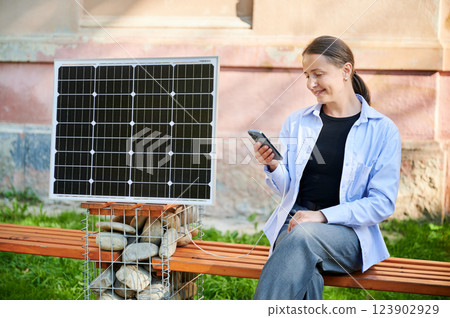 Happy woman using smartphone connected to photovoltaic solar panel. Integration of sustainable renewable energy into everyday life, demonstrating practical use of solar power for charging devices. 123902929