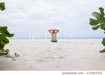 A young woman is sitting on the sand on the beach and enjoying the beautiful seascape. A girl in a swimsuit and a hat is relaxing by the ocean. Back view. 123903501