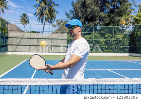 A young man in a cap and sunglasses throws a yellow ball and holds a racket. An athlete plays pickleball outdoors. A young man in a cap and sunglasses throws a yellow ball and holds a racket. An athlete plays pickleball outdoors. 123903509