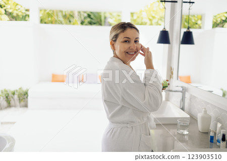 A young woman in a white bathrobe applies cream to her neck in the bathroom A young woman in a white bathrobe applies cream to her neck in the bathroom 123903510