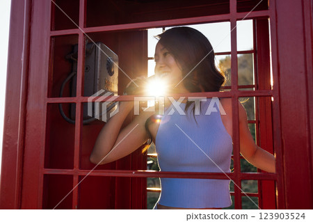 An attractive Asian woman in casual clothes is talking on an old phone in a red outdoor phone booth. A happy charming Korean woman laughs and calls. 123903524