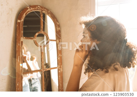 A close-up of a charming mixed-race teenager puts a cosmetic cloth mask on her face. The reflection of an attractive African girl in the mirror. 123903530