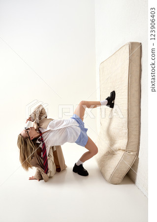 Full-length dynamic pose of young woman posing bending backward, leg resting on mattress, styled in schoolgirl-inspired business attire. 123906403