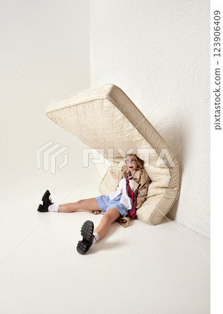 Playful image of young woman sitting under mattress with exaggerated expression, wearing trench coat and loose tie. 123906409