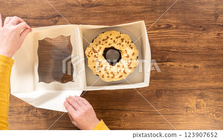 Flat lay. Carefully placing the gingerbread bundt cake, adorned with salted caramel frosting and gingerbread sprinkles, into a white paper bundt cake box with a clear window for gifting. 123907652