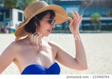 A woman in a blue swimsuit and straw hat stands on the stunning beach of Dubai, gazing at the impressive skyline filled with modern skyscrapers, while the golden sand and gentle waves create a perfect 123908764
