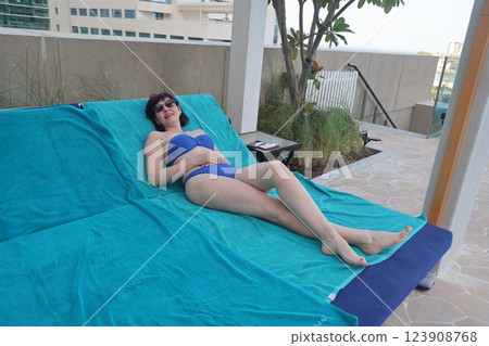 Relaxing by the Pool: A Sun-Kissed Woman in a Bikini and Sunglasses Enjoys the Warm Summer Day on a Blue Towel, Embracing the Joy of Leisure and Tranquility Under the Clear Sky, Surrounded by the 123908768