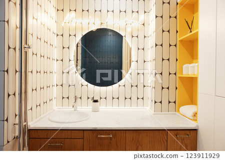 Modern stylish bathroom interior. Close-up of marble countertop with sink and large round illuminated mirror. Bright accent on yellow shelves with household items. Modern stylish bathroom interior. Close-up of marble countertop with sink and large round illuminated mirror. Bright accent on yellow shelves with household items. 123911929