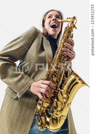 Emotional and joyful young woman, saxophone player in stylish blazer playing with energy against white background. 123912155