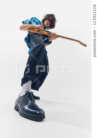 Low-angle image of stylish young man, guitarist in wide stance, dynamically playing guitar against white background. 123912228