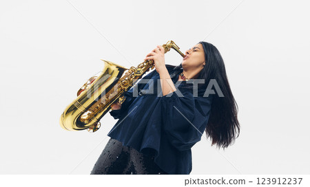 Woman in dark loose clothing passionately playing saxophone, eyes closed, fully immersed in rhythm, isolated on white background. 123912237