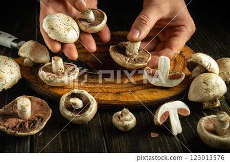 Two hands hold and slice various mushrooms on a wooden cutting board. The fresh mushrooms are being prepared for cooking, showcasing their textures and earthy colors 123913576