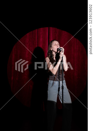 Young woman in jeans and a T-shirt standing behind a microphone on stage and singing. Performing a monologue on stage Young woman in jeans and a T-shirt standing behind a microphone on stage and singing. Performing a monologue on stage 123913602