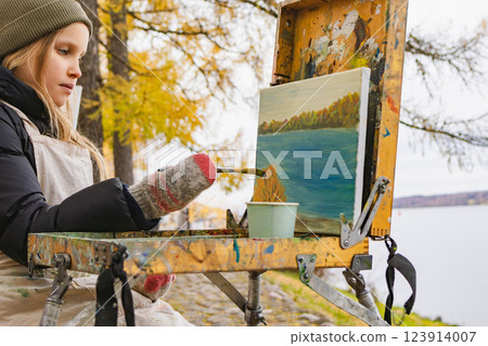 Young artist painting a landscape outdoors near a river on a cold autumn day 123914007