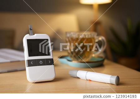 A glucometer sits on a wooden table next to a cup of tea. The cozy indoor atmosphere is enhanced by soft lighting and a nearby plant, indicating a moment of self-care A glucometer sits on a wooden table next to a cup of tea. The cozy indoor atmosphere is enhanced by soft lighting and a nearby plant, indicating a moment of self-care 123914193