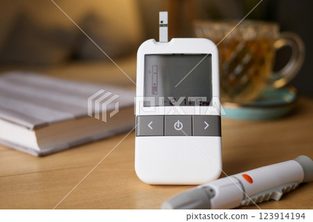 A glucometer sits on a wooden table, displaying the time and ready for a blood sugar test. Nearby, a cup of tea and an opened notebook add to the setting A glucometer sits on a wooden table, displaying the time and ready for a blood sugar test. Nearby, a cup of tea and an opened notebook add to the setting 123914194