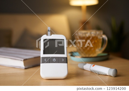 A glucometer is placed on a wooden table, showing blood sugar test results. Nearby, a cup of tea and a pen for logging data create a calming atmosphere in a cozy living space 123914200