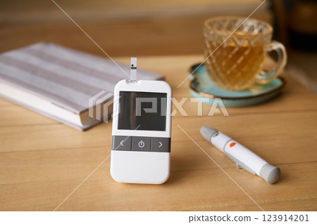 A glucometer sits on a wooden table next to a notebook and a cup of tea. A pen lies nearby, indicating a moment of personal health monitoring at home A glucometer sits on a wooden table next to a notebook and a cup of tea. A pen lies nearby, indicating a moment of personal health monitoring at home 123914201