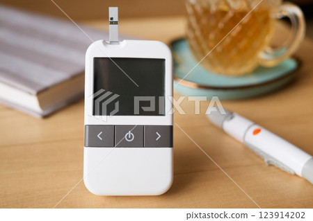 A glucometer rests on a wooden table beside a drink, ready for a blood sugar test. A pen for insulin dosage is also visible, indicating health management focus 123914202