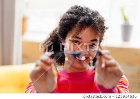 A young African woman smiles and keeps her glasses at home. A short-sighted attractive black girl laughs and wipes her glasses indoors. 123914636