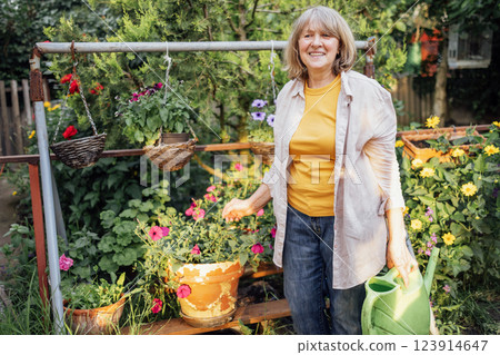 An elderly smiling woman waters flowers in hanging pots from a watering can 123914647