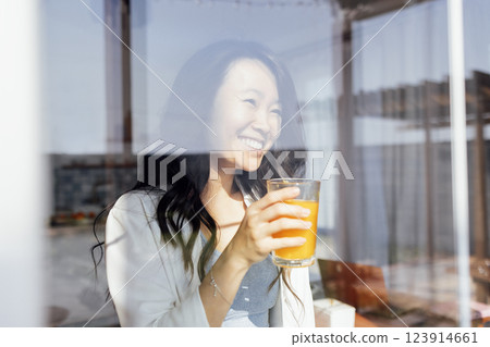 An attractive young Asian woman holds a glass of orange juice and laughs. Reflection in the window. 123914661