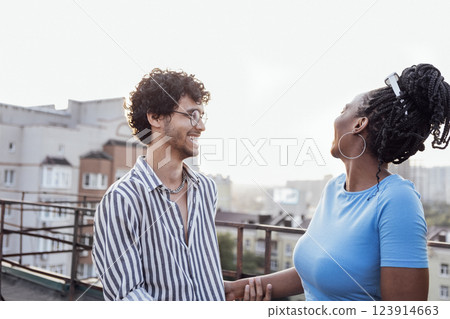 Young happy couple in love on roof of multi-storey building. Cute curly-haired interracial guy holds hand of charming African woman. 123914663