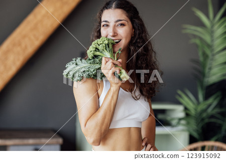 An attractive smiling vegan girl in a white top bites fresh broccoli. A curly-haired young fit woman laughs and eats cabbage at home. An attractive smiling vegan girl in a white top bites fresh broccoli. A curly-haired young fit woman laughs and eats cabbage at home. 123915092