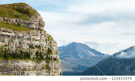 A rugged, rocky cliff face rises sharply from a valley floor. Lush green vegetation covers the cliff face, while a mountain range with forested slopes and cloudy peaks rises in the background A rugged, rocky cliff face rises sharply from a valley floor. Lush green vegetation covers the cliff face, while a mountain range with forested slopes and cloudy peaks rises in the background 123915474