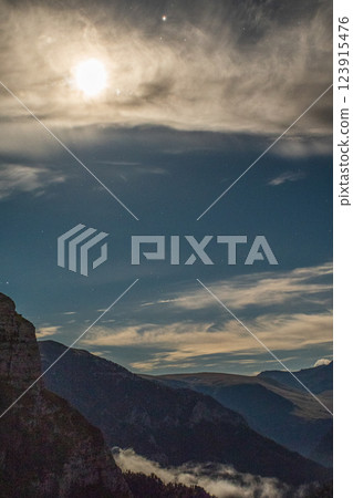 A moonlit landscape with mountains, clouds, and a bright moon. Stars are visible in the night sky 123915476
