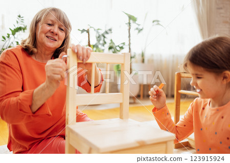 Elderly woman in casual clothes and her little granddaughter are assembling new wooden chair Elderly woman in casual clothes and her little granddaughter are assembling new wooden chair 123915924