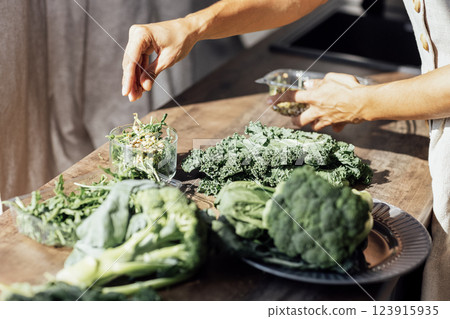 Close-up of a female hands making a green salad in the kitchen. A young woman pours pine nuts into a glass bowl with arugula, broccoli and curly cabbage leaves. Close-up of a female hands making a green salad in the kitchen. A young woman pours pine nuts into a glass bowl with arugula, broccoli and curly cabbage leaves. 123915935