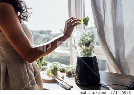 A close-up of a blender with green vegetables and herbs and a female hands with a leaf of wavy cabbage. The girl makes a vitamin smoothie at home. A close-up of a blender with green vegetables and herbs and a female hands with a leaf of wavy cabbage. The girl makes a vitamin smoothie at home. 123915936