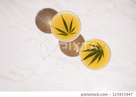 Two elegant long-stemmed glasses with green cocktails with cannabis leaves on the light marble surface of the table. A delicious healthy drink with hemp on a white background. 123915947